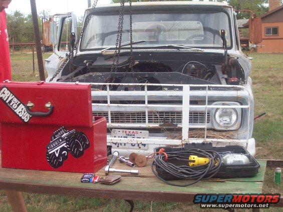 Old pickup truck with hood open, chains attached to engine bay. Red toolbox with stickers on table in front