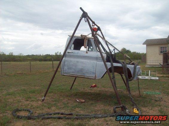 Gray truck cab, stripped down and suspended by a hoist, sits in a grassy yard