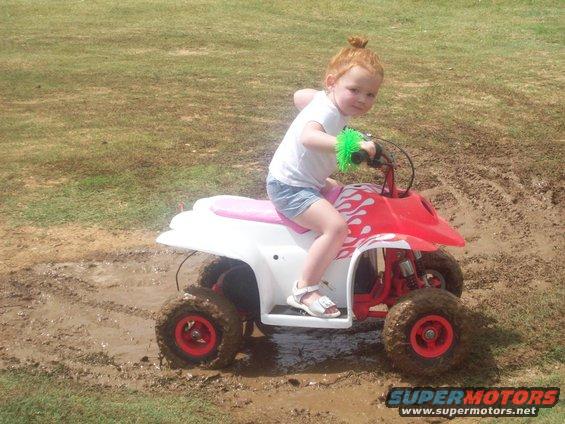 Child with red hair rides a red and white ATV through mud. The ATV has flame decals