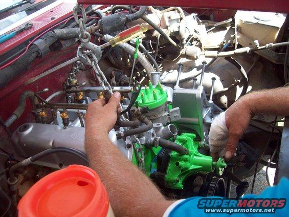 Engine bay with green painted parts, chain hoist, and hands working on it. Supermotors.net logo visible