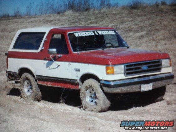 Red and white Ford Bronco stuck in dirt, front 3/4 view. Windshield banner reads 'WEEKEND 4 WHEELERS'