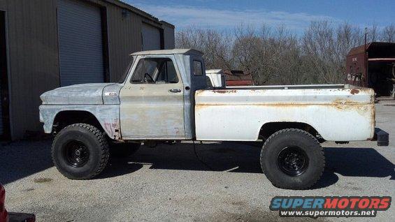 Old pickup truck with big tires, primer on front, white bed. Parked outside a garage
