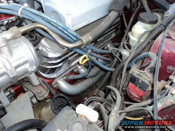 Engine bay with hoses, wires, and engine components visible. Some text on a red and black part