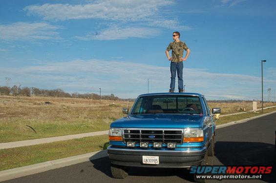 Blue 1992 Ford F-150 with aftermarket lights on the grille, license plate LN30985, and a person standing on its roof
