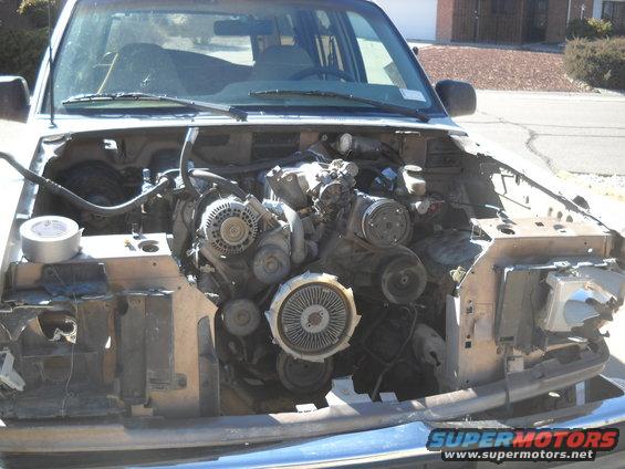 Vehicle engine bay with engine exposed, front end partially dismantled. Duct tape is visible on the left
