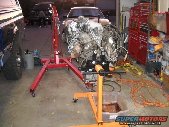 Engine on an orange engine stand, with a red engine hoist nearby. A pickup truck is partially visible on the left