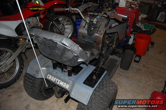 Riding mower with a gray body and a taped-up seat, parked next to a dirt bike
