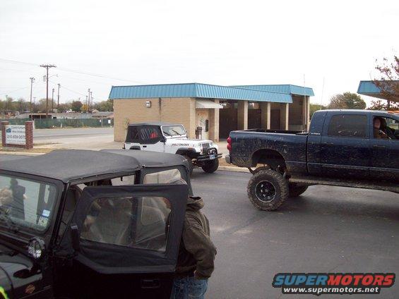 Three lifted off-road vehicles parked near a building with a blue roof. One is a black Jeep in the foreground, another is a white Jeep, and the third is a dark blue pickup truck