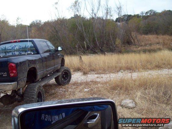 Lifted black pickup truck with large tires parked on a dirt path next to dry grass and trees. Side mirror reflects a blue shirt
