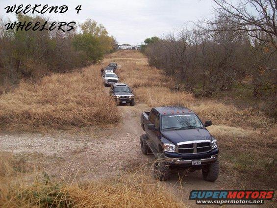 Dark blue pickup truck leads a line of SUVs on a dirt path through dry grass and trees