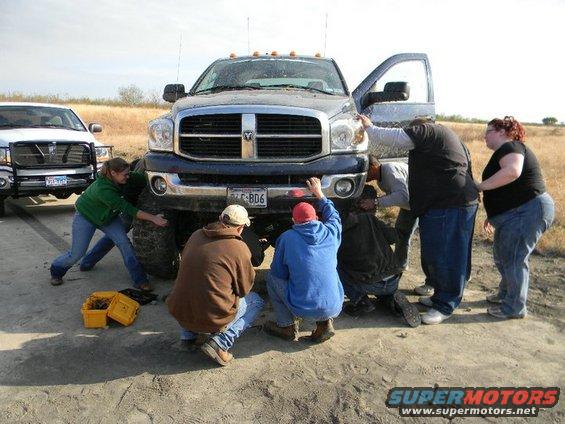 Front view of dark blue Dodge Ram truck with people working on it; white truck in background