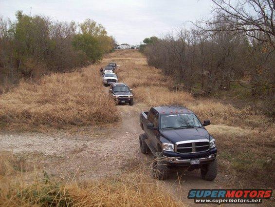 Line of SUVs and trucks driving on a dirt road through dry grass and trees