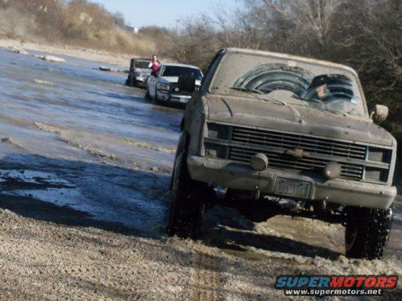 Muddy pickup truck driving through water, with other trucks behind it