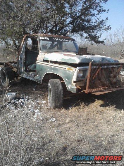 Rusty green and white Ford F-Series truck with a damaged front end and missing doors, parked in a dry, grassy field