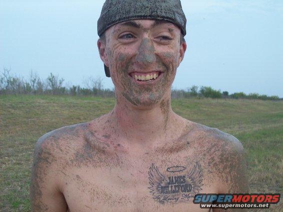 Mud-covered person wearing a cap smiles broadly, with a tattoo on chest