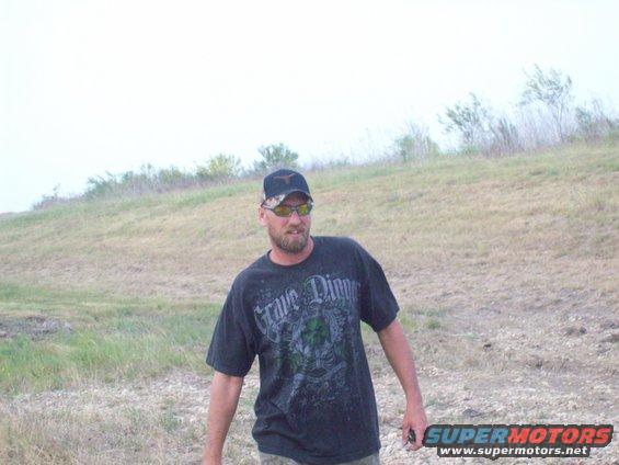 Man wearing a black t-shirt with 'Grave Digger' graphic and a Texas Longhorns cap stands in a grassy field