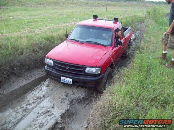 Red pickup truck with license plate BJ5006N stuck in a muddy ditch, driver visible inside