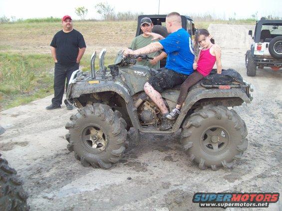 Muddy ATV with two adults and a child riding, two other people standing nearby, and a Jeep in the background