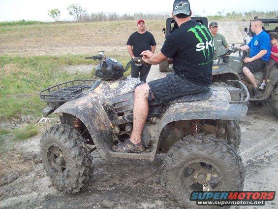 Mud-covered ATV with camouflage pattern, side view. Rider in black shorts and Monster Energy shirt