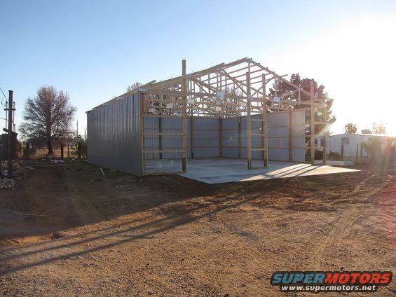 Metal building under construction with wooden roof trusses and corrugated metal siding. Dirt ground in foreground