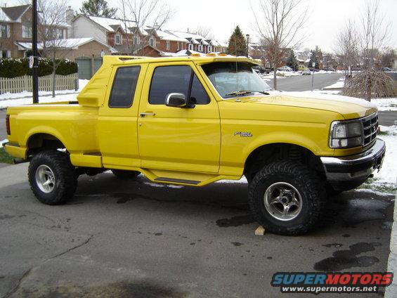 Yellow Ford F-150 pickup truck with custom bed cap, lifted suspension, and aftermarket wheels, parked on a residential street