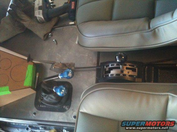 Vehicle interior showing two gear shifters, one with blue knobs, and parts of two seats. A power tool is visible in the background