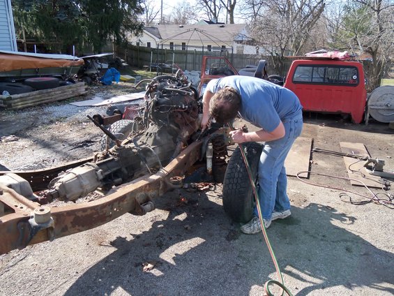 Man bends over a rusty truck chassis with engine removed. Red truck cab in background