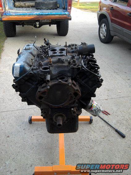 Engine on an orange stand, with a blue pickup truck and a red SUV in the background