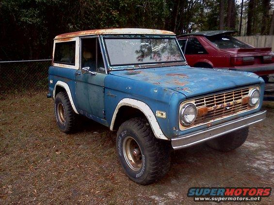 Blue 1974 Ford Bronco with white hardtop parked outdoors, red car behind it