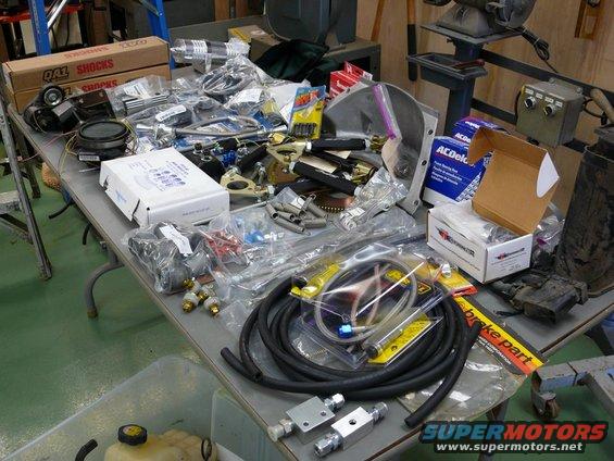 Table covered in car parts like shocks, hoses, and engine components. Some boxes have brand names like QA1 and ACDelco