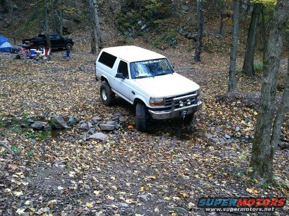 White SUV with lifted suspension parked in a forest campsite. Another black truck is visible in the background