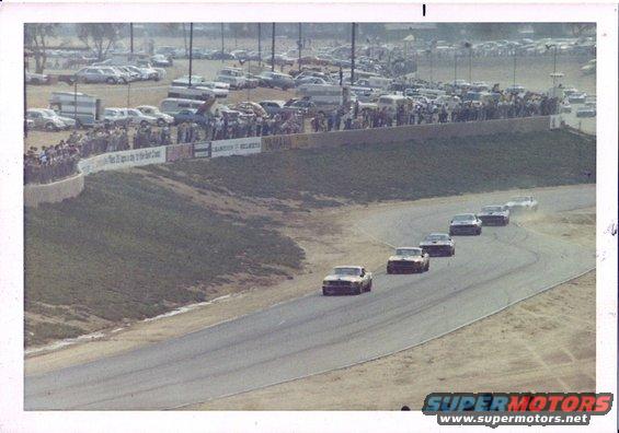 Several race cars speed around a track with spectators in the background