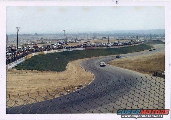 Race cars on a track with spectators and parked cars in the background