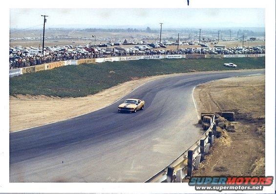 Yellow 1970 Ford Mustang race car on track, front view, with spectators and parked cars in background