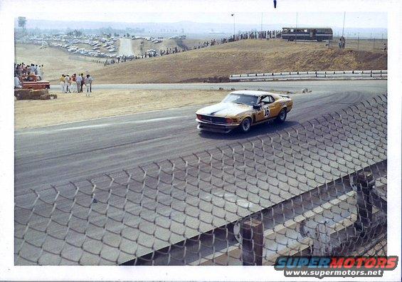 Yellow race car with black stripes and number 16 on side, turning on a track with spectators and parked cars in background