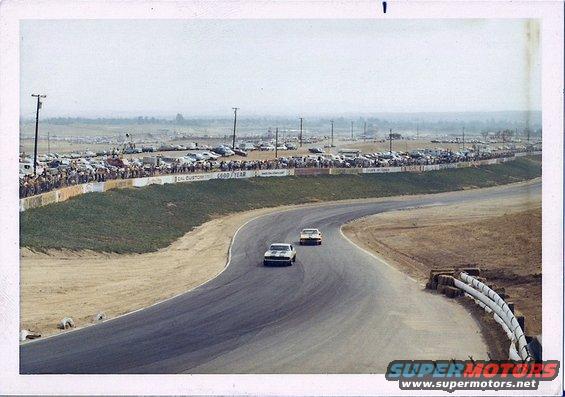 1973 Ford Mustang race cars on track, spectators in background, many parked cars