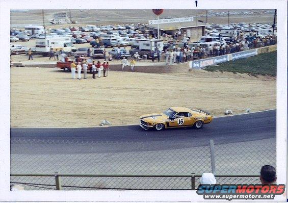 Yellow 1970 Ford Mustang Boss 302 race car with black stripes and number 16 on the door, seen from the front-right at a racetrack