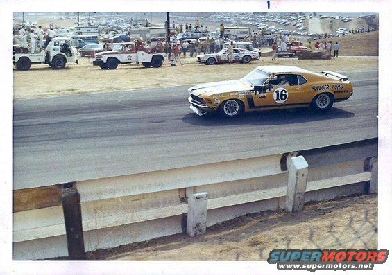 Yellow race car with number 16 and 'FOULGER FORD' text on track, other cars and tow trucks in background