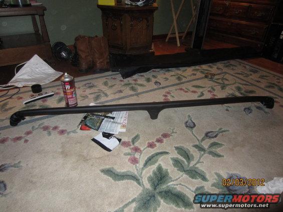 Black car part, possibly a roof rack or trim piece, lying on a floral rug next to a can of spray paint and a remote
