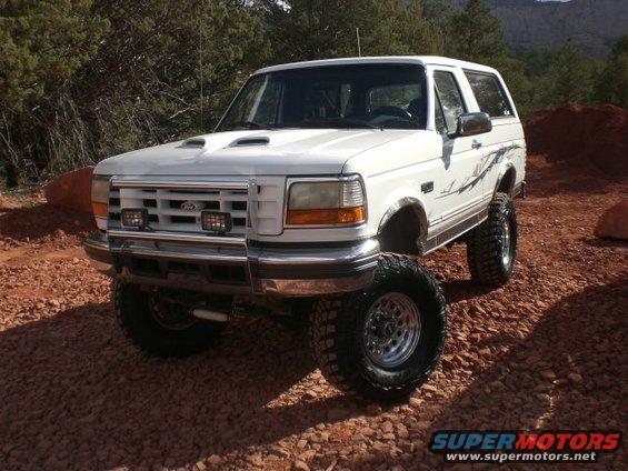 White 1996 Ford Bronco lifted with large off-road tires, parked on gravel with trees and hills in background