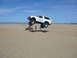 1988 Ford Bronco - A white 1988 Ford Bronco soars through the air during a jump over sand dunes, with an ATV and onlookers below.