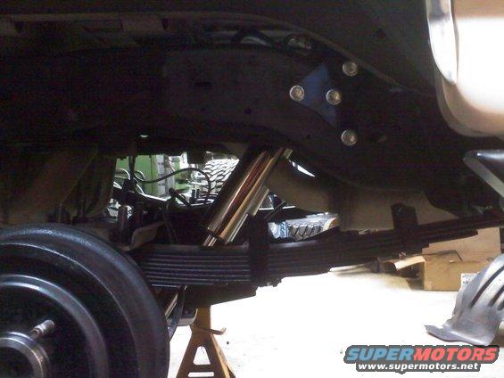 Undercarriage view showing leaf springs, a shock absorber, and part of a wheel. A blue bracket is bolted to the frame