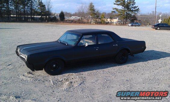 Black 1966 Oldsmobile Cutlass sedan parked on gravel lot, matte finish, black wheels