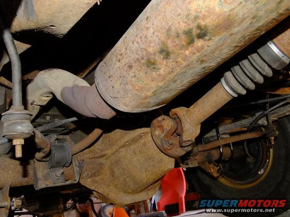 Rusty rear differential and exhaust pipe under a vehicle. Visible driveshaft and suspension components
