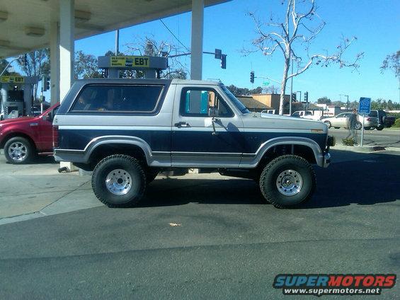 Two-tone Ford Bronco with lifted suspension and off-road tires parked at a gas station. Red SUV is also visible