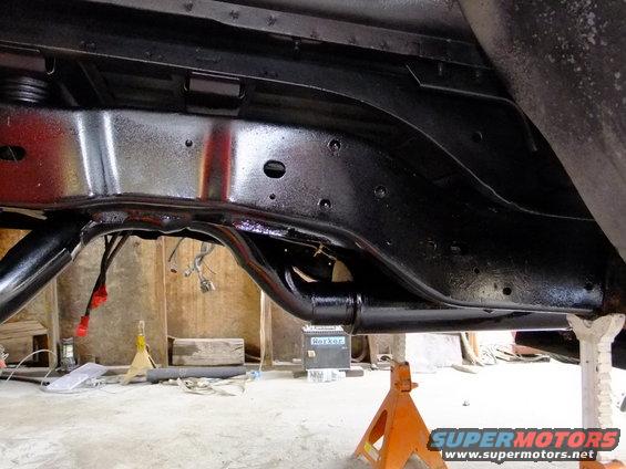 Underside of a vehicle frame, painted black, supported by jack stands. Some wiring is visible