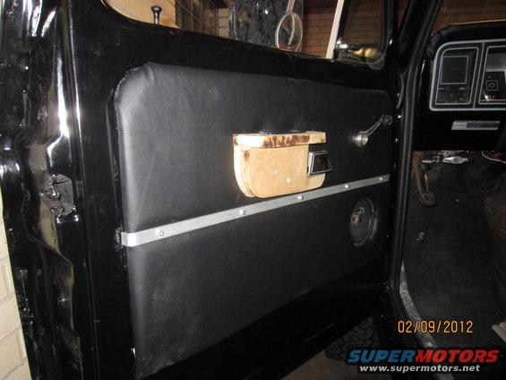 Black interior door panel with chrome handle, wooden armrest, and speaker grille. Dashboard visible