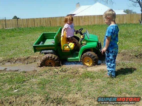 Child driving a green toy John Deere Gator stuck in mud, another child watches