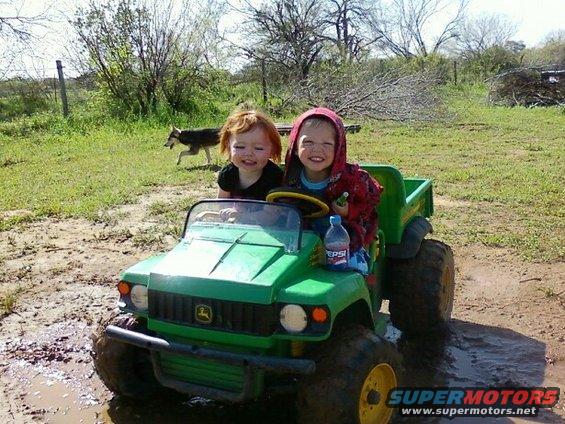 Two kids in a green toy tractor driving through mud. A dog runs in the background