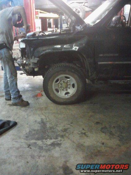 Mechanic works on the damaged front fender of a black Chevrolet 2500 HD truck. The hood is open